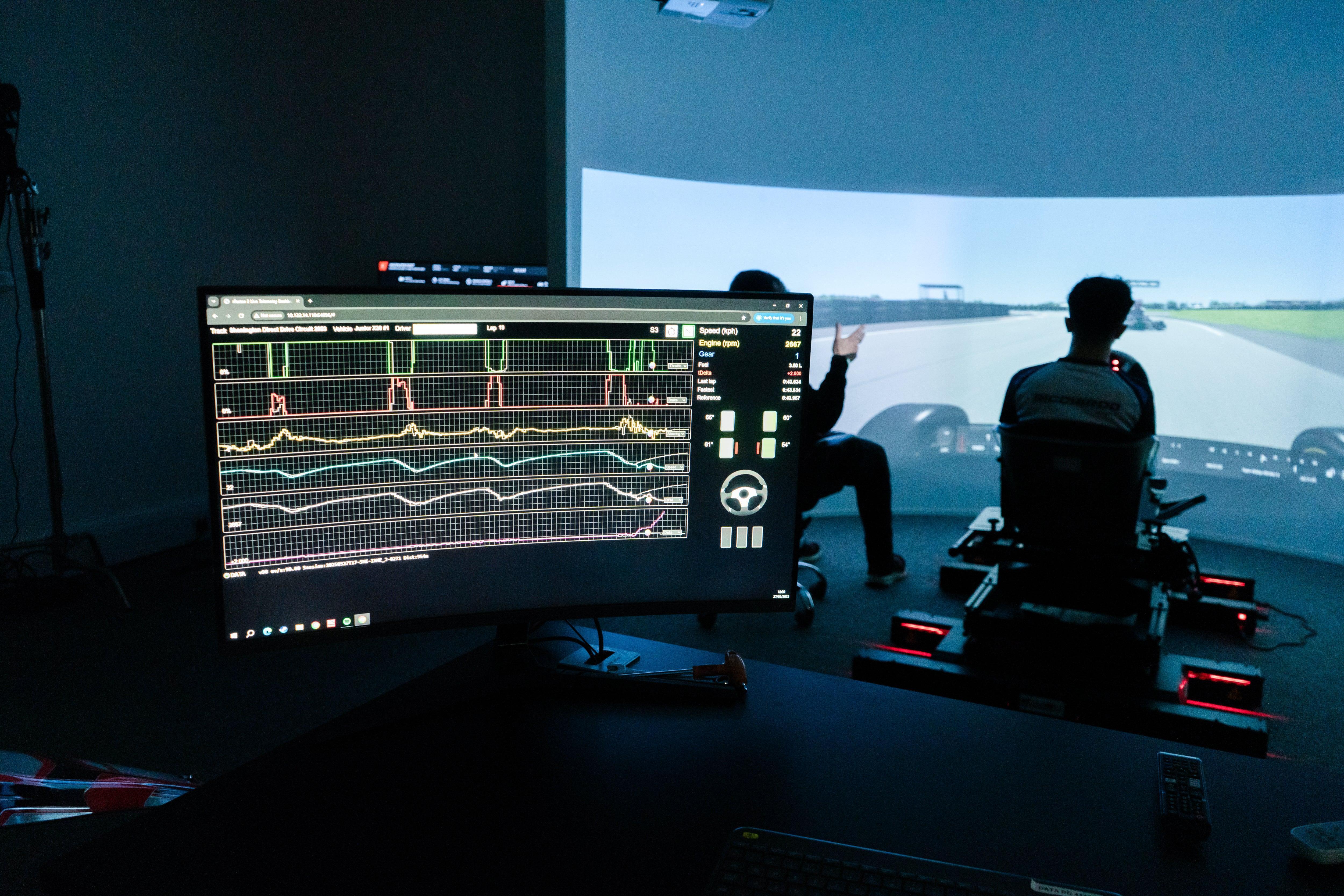 A KartSim driver coaching session showing a large curved racing simulator screen with a driver in the seat, while a coach points at the track view; a foreground monitor displays telemetry data and performance graphs for analysis.