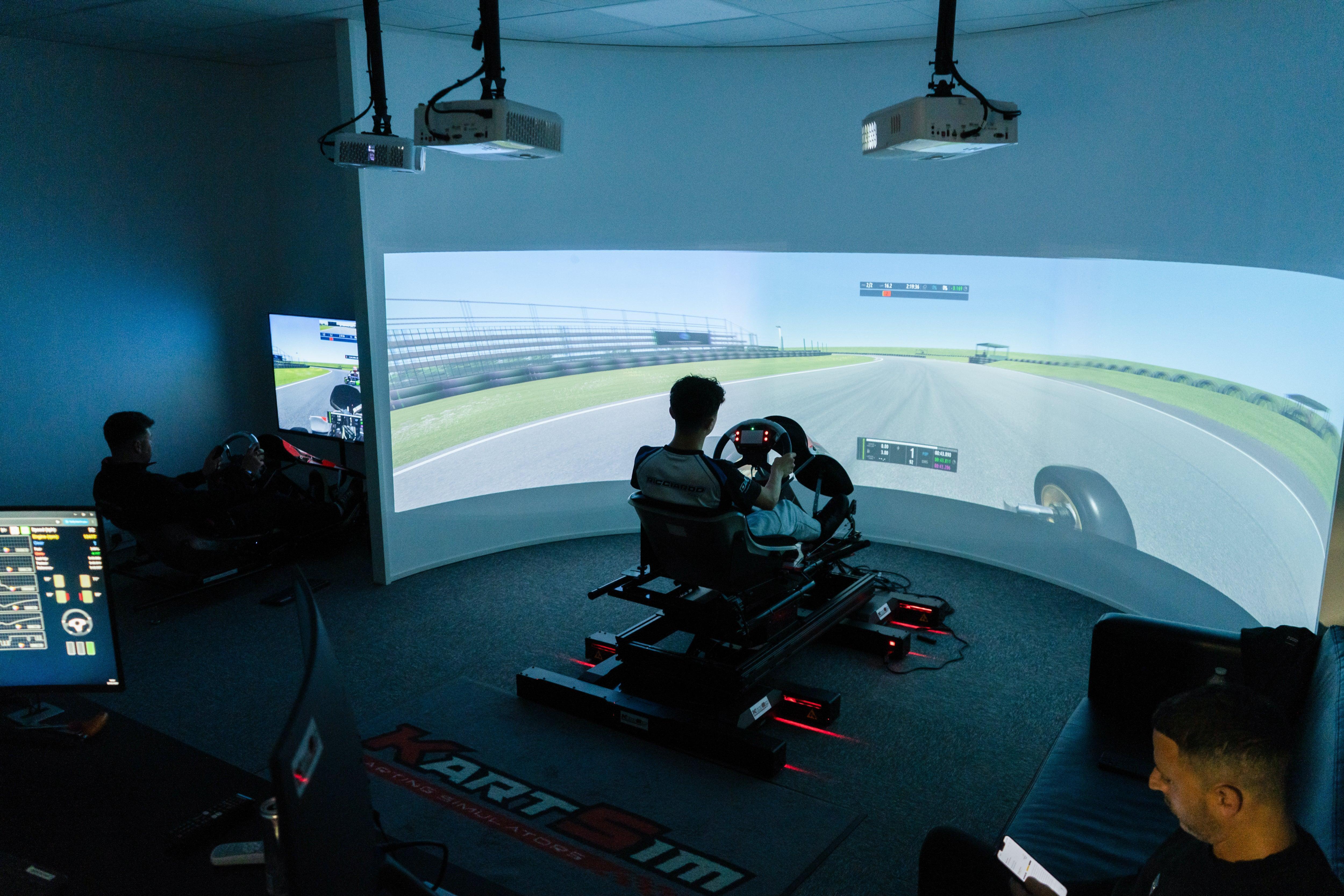 A KartSim driver coaching session showing a large curved racing simulator screen with a driver in the seat, while a coach points at the track view; a foreground monitor displays telemetry data and performance graphs for analysis.