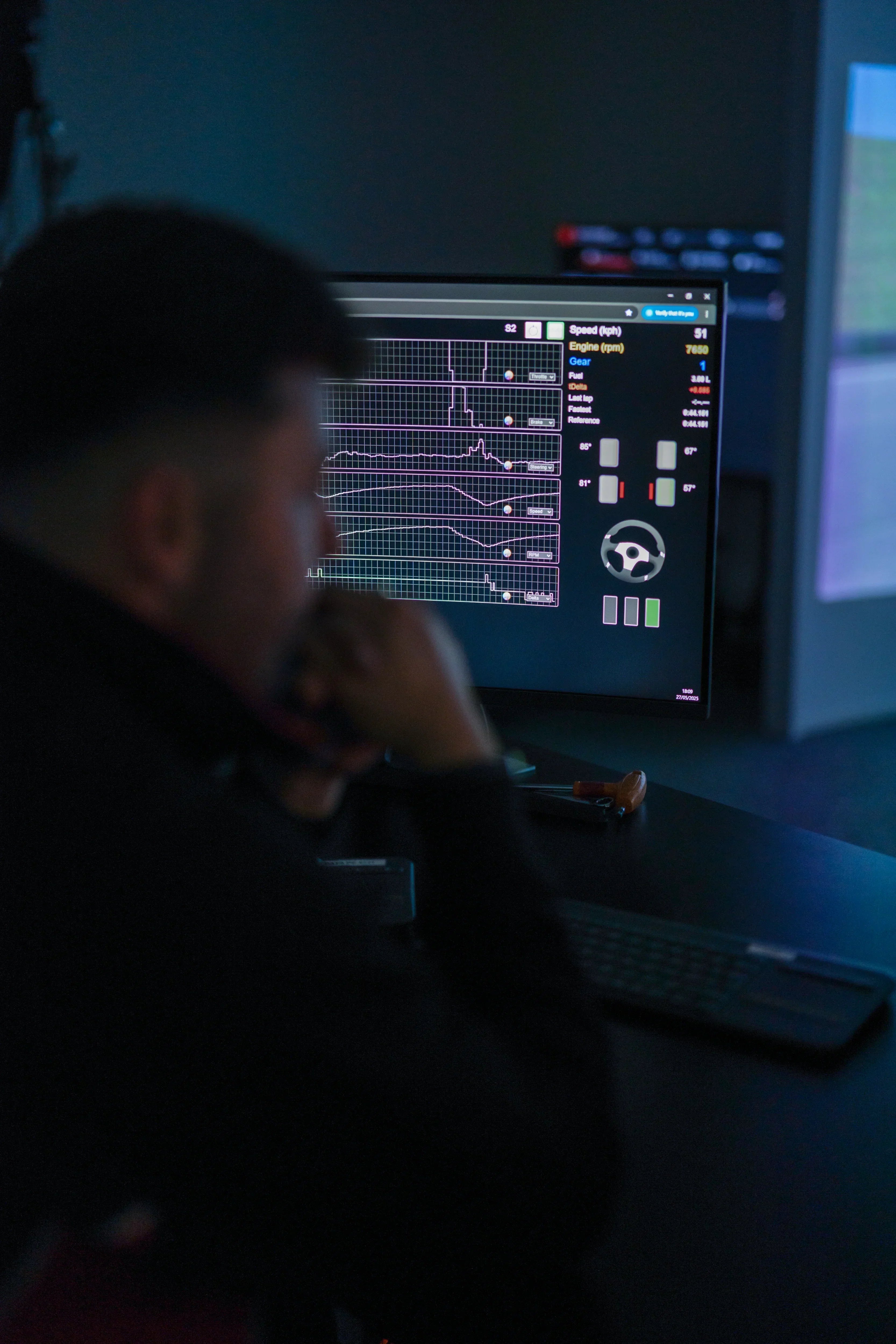 A KartSim driver coaching session showing a large curved racing simulator screen with a driver in the seat, while a coach points at the track view; a foreground monitor displays telemetry data and performance graphs for analysis.