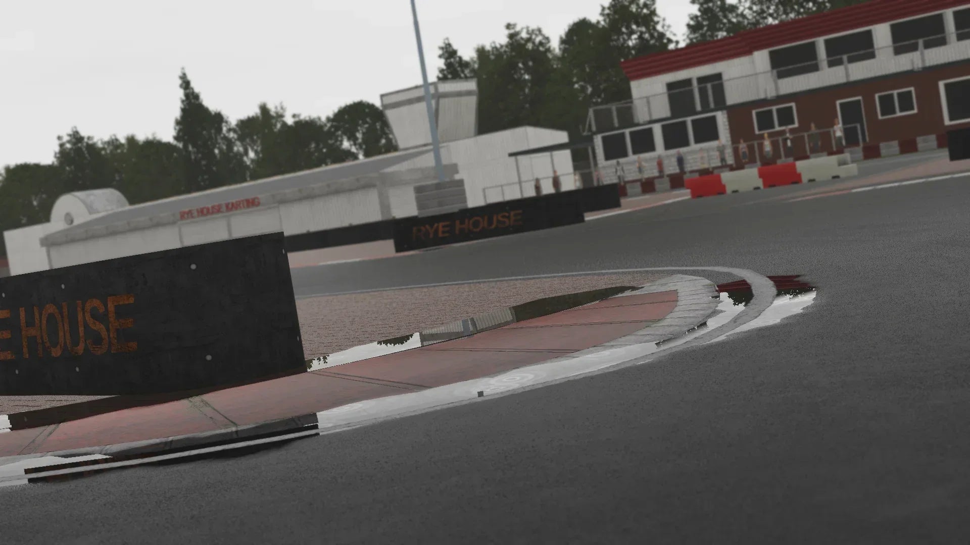 Close-up of a wet curb and puddle reflecting the “Rye House” branding at KartSim’s Rye House track, with the karting building in the background.
