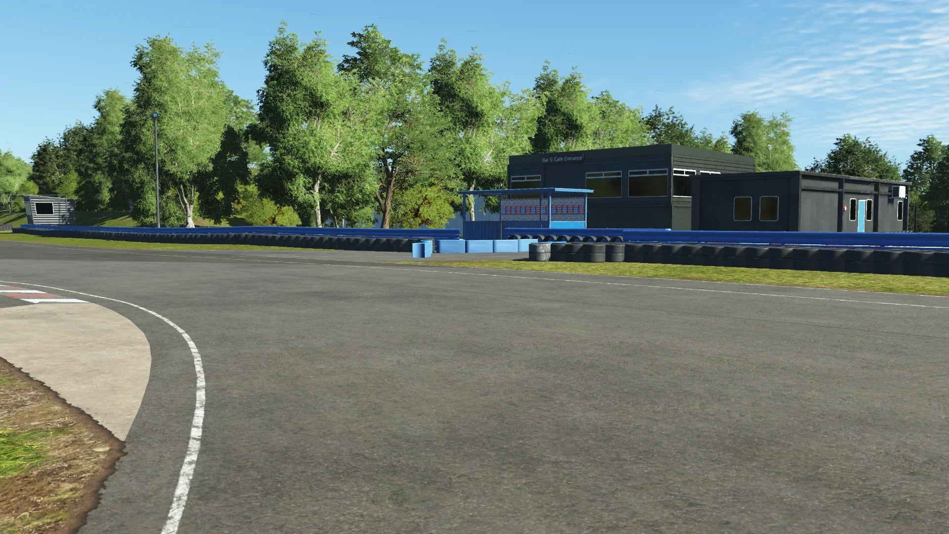 Trackside view of Teesside in KartSim, focusing on a corner near the main building with blue barriers and a race control tower, set against a backdrop of lush green trees under clear skies.