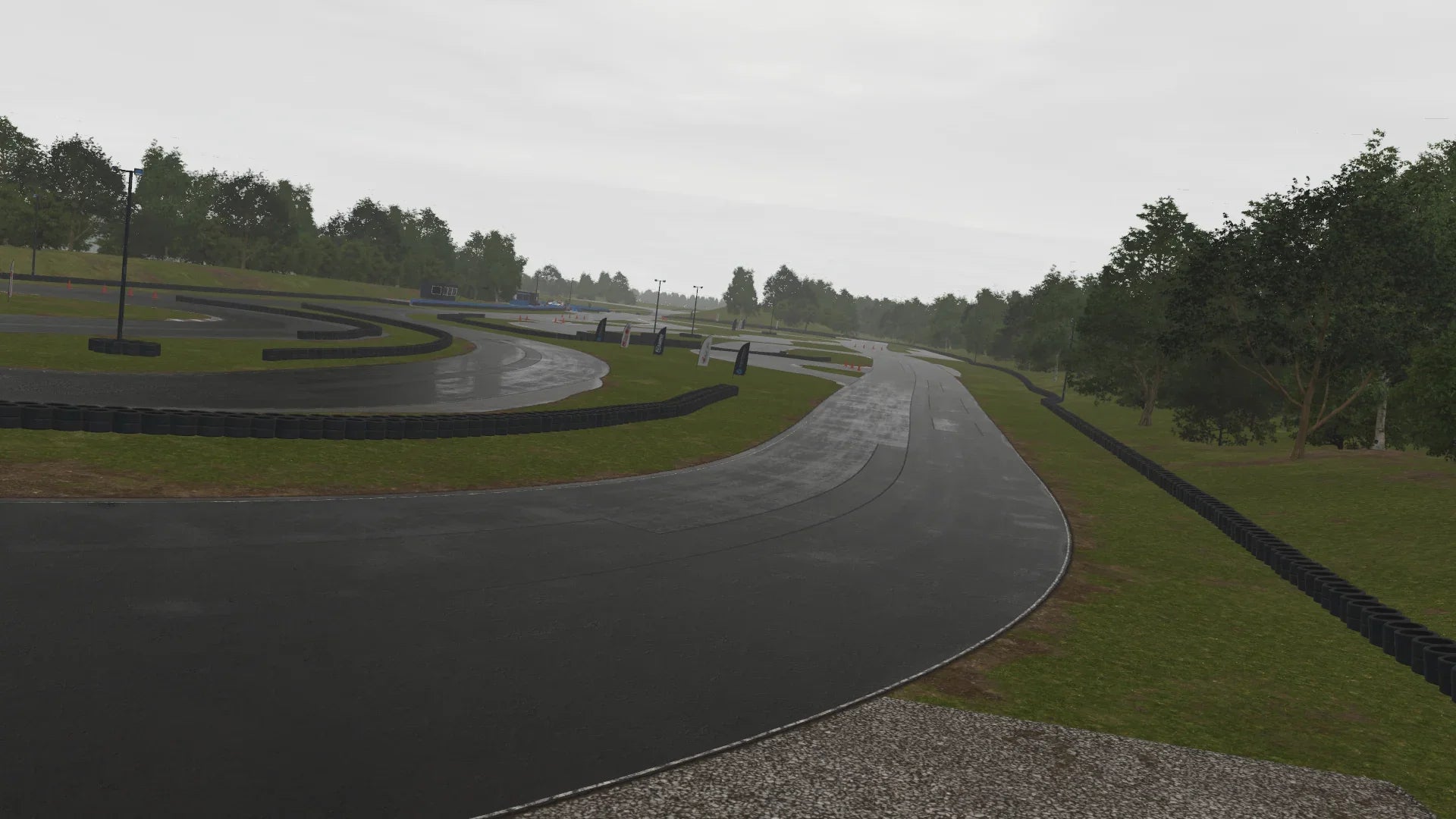 Overcast view of Teesside in KartSim, showing a sharp black asphalt corner bordered by grassy banks and tyre barriers, with trees swaying gently in the background.