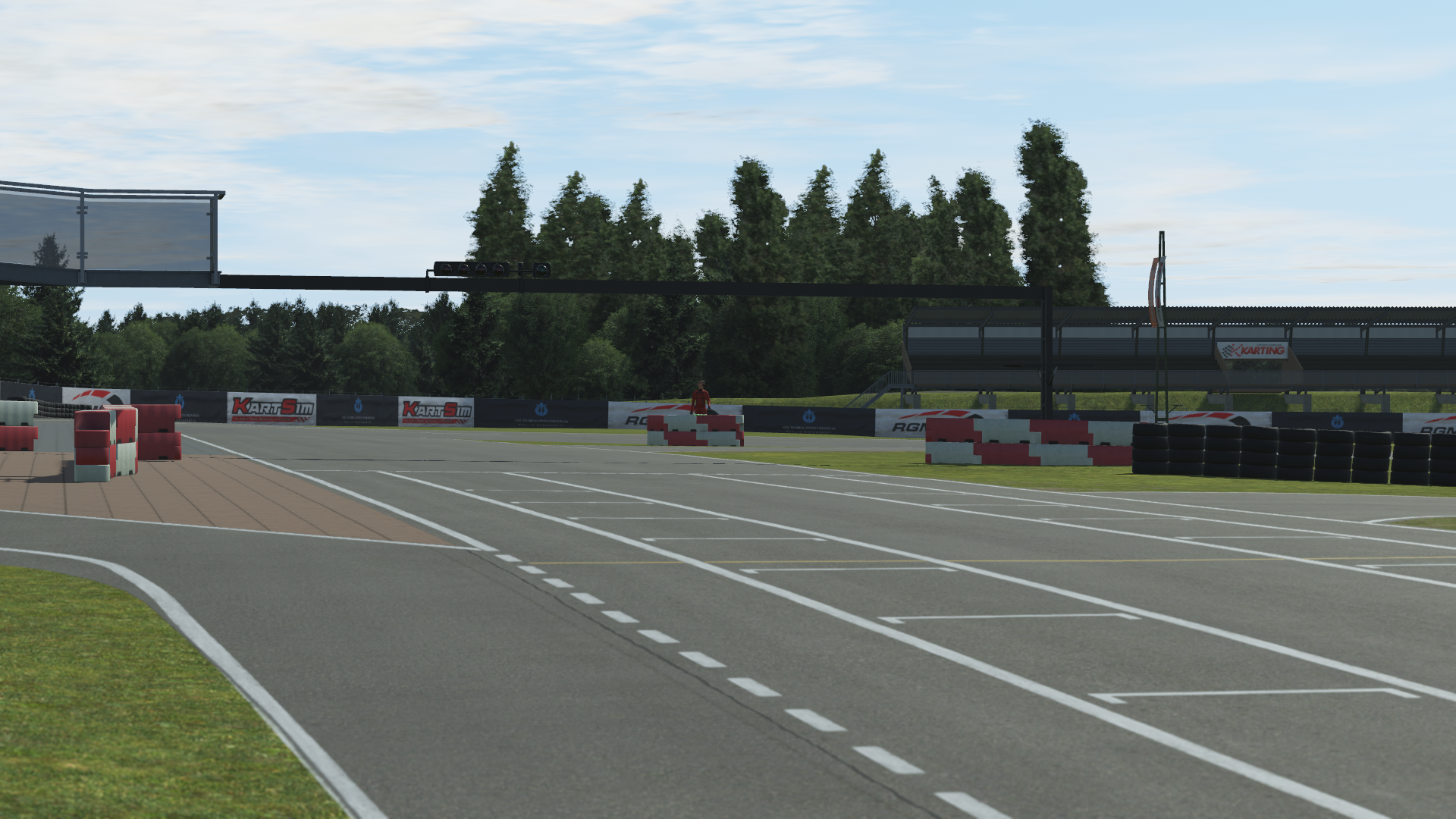 KartSim software screenshot of the Kristianstad circuit start grid: white grid markings on smooth asphalt, red-and-white tire barriers and stacks lining the track edge, an overhead gantry with signal lights, and a row of tall trees in the background under a blue sky.