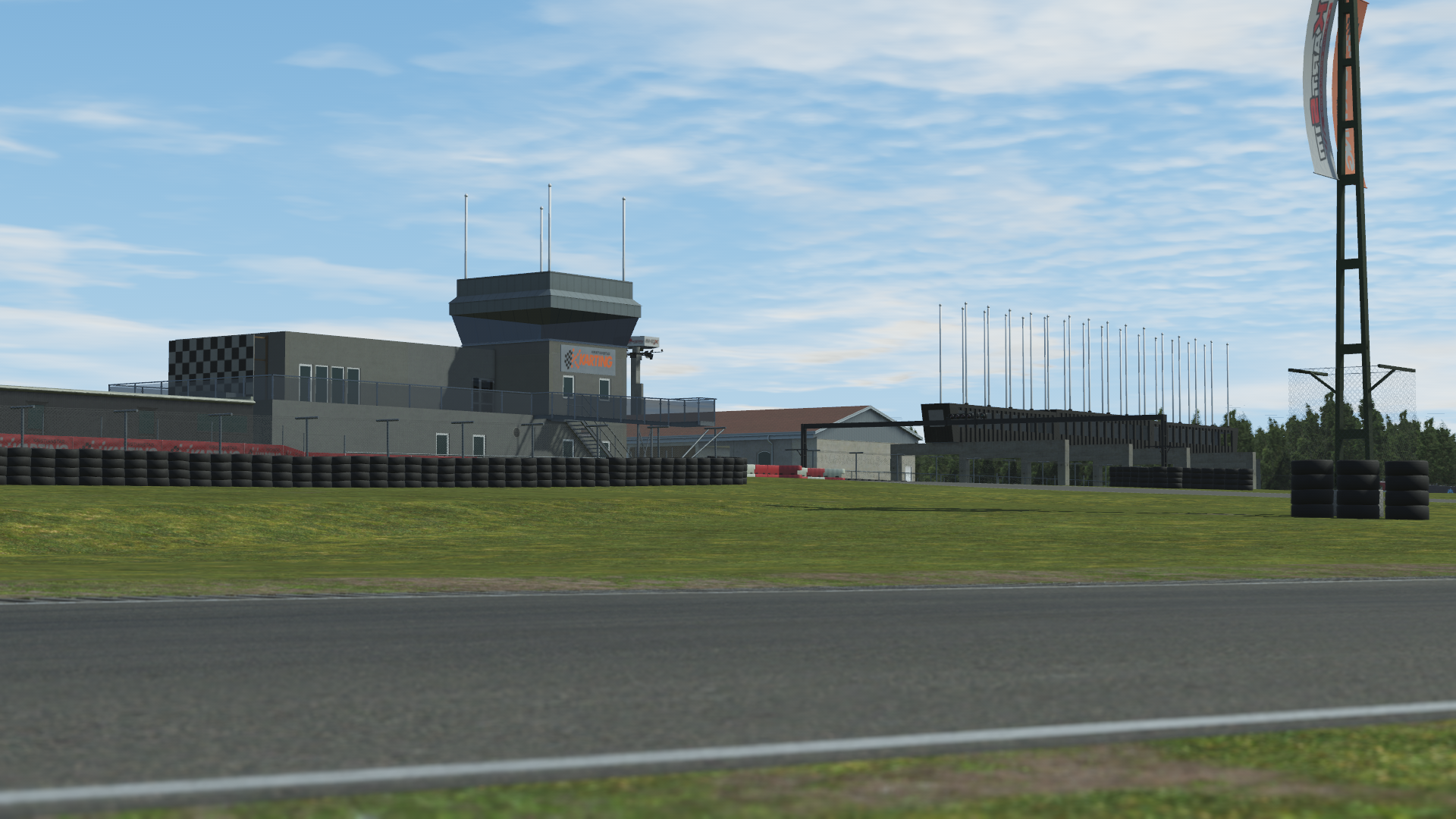 KartSim software rendering of the Kristianstad circuit pit complex: low-angle view showing the asphalt track edge in the foreground, green grass verge, stacked black tire barriers, a grey pit building with checkered livery and adjacent control tower, rows of empty flagpoles, and a tall K artSim-branded tower under a clear blue sky.