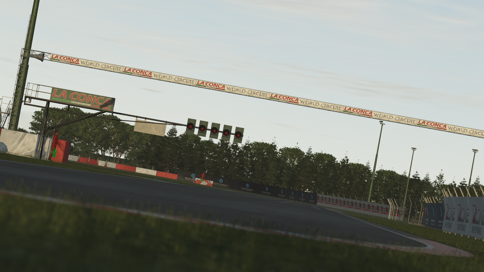 KartSim software render of the La Conca World Circuit’s start/finish straight, featuring the overhead “La Conca World Circuit” arch, a row of signal lights, red-and-white armco barriers, and a backdrop of dense trees under a pale sky.