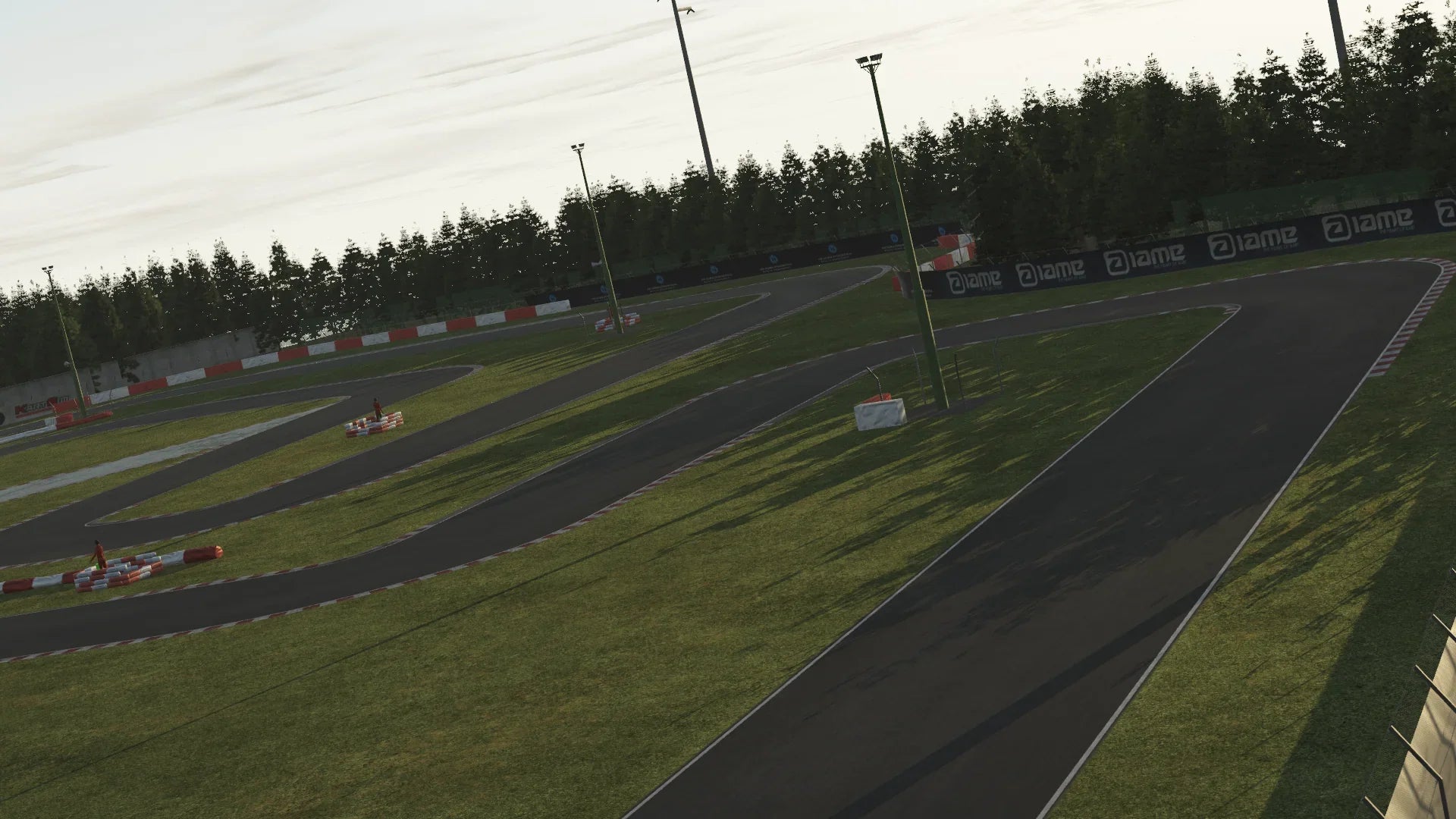 KartSim software render of the La Conca kart track at dusk, showing a sweeping asphalt curve alongside green grass, red-and-white safety barriers, tall floodlight poles, and dense pine trees under a pale evening sky.