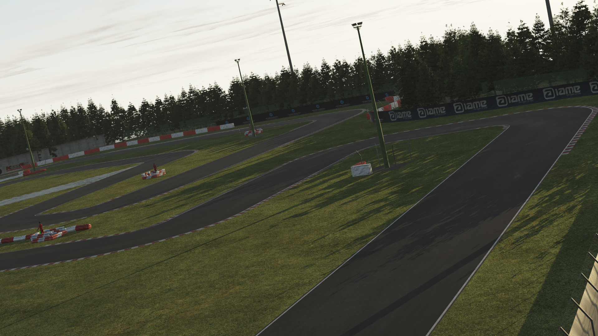 KartSim software render of the La Conca kart track at dusk, showing a sweeping asphalt curve alongside green grass, red-and-white safety barriers, tall floodlight poles, and dense pine trees under a pale evening sky.