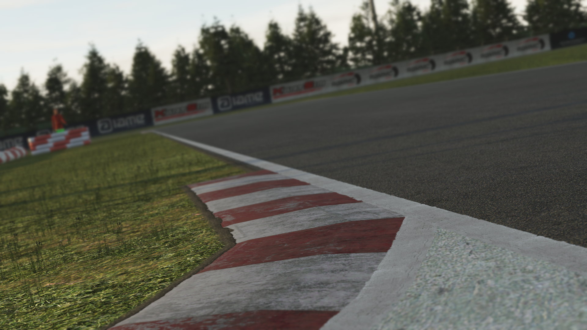 KartSim software render showing a close-up of the La Conca kart track’s red-and-white striped kerb beside dark asphalt, bordered by a grassy verge and lined with sponsorship banners in front of a row of pine trees under a dusky sky.
