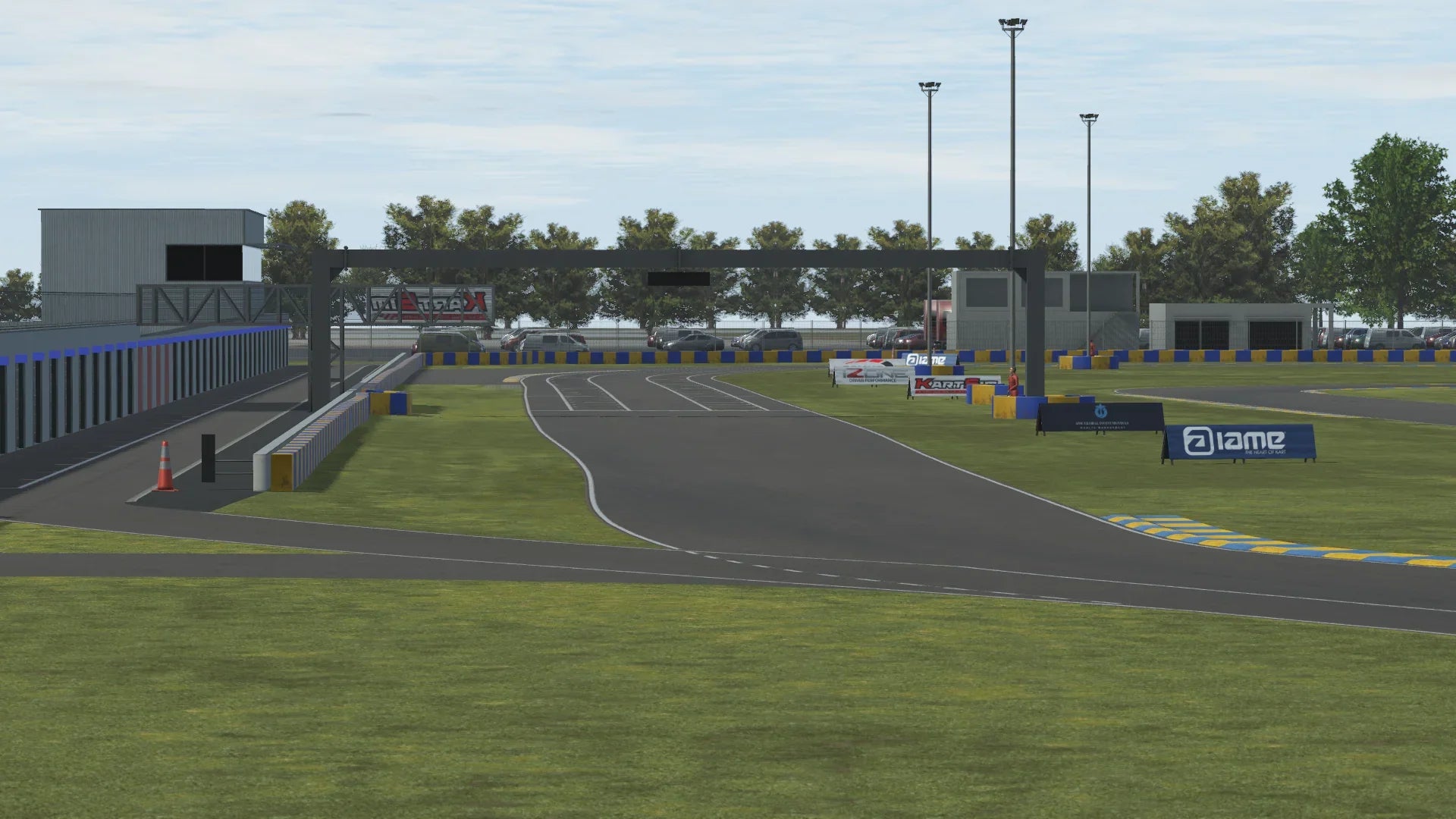 KartSim software screenshot of a Le Mans–style kart track showing the pit lane exit merging onto the main straight, bordered by green grass, blue-and-yellow kerbs, marshal posts, advertising banners, light poles, a pit building on the left, parked cars and trees in the background under a partly cloudy sky.