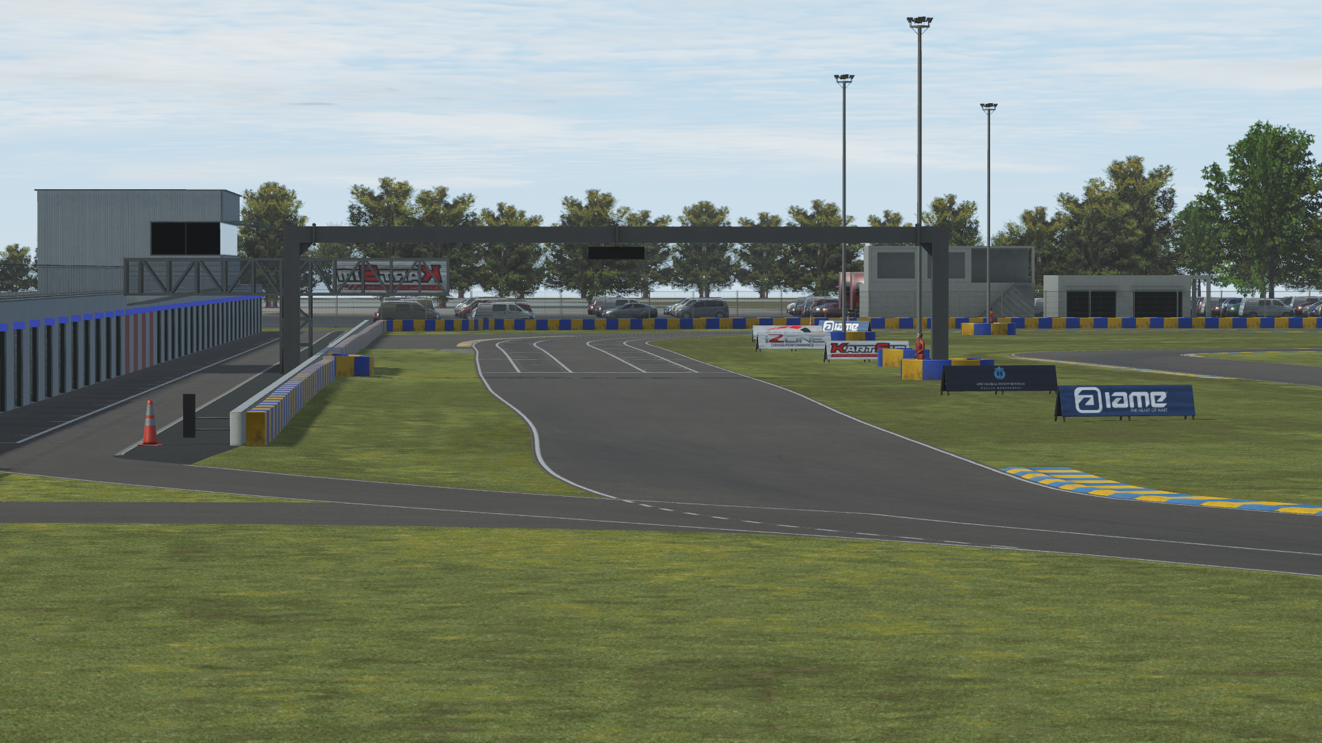 KartSim software screenshot of a Le Mans–style kart track showing the pit lane exit merging onto the main straight, bordered by green grass, blue-and-yellow kerbs, marshal posts, advertising banners, light poles, a pit building on the left, parked cars and trees in the background under a partly cloudy sky.