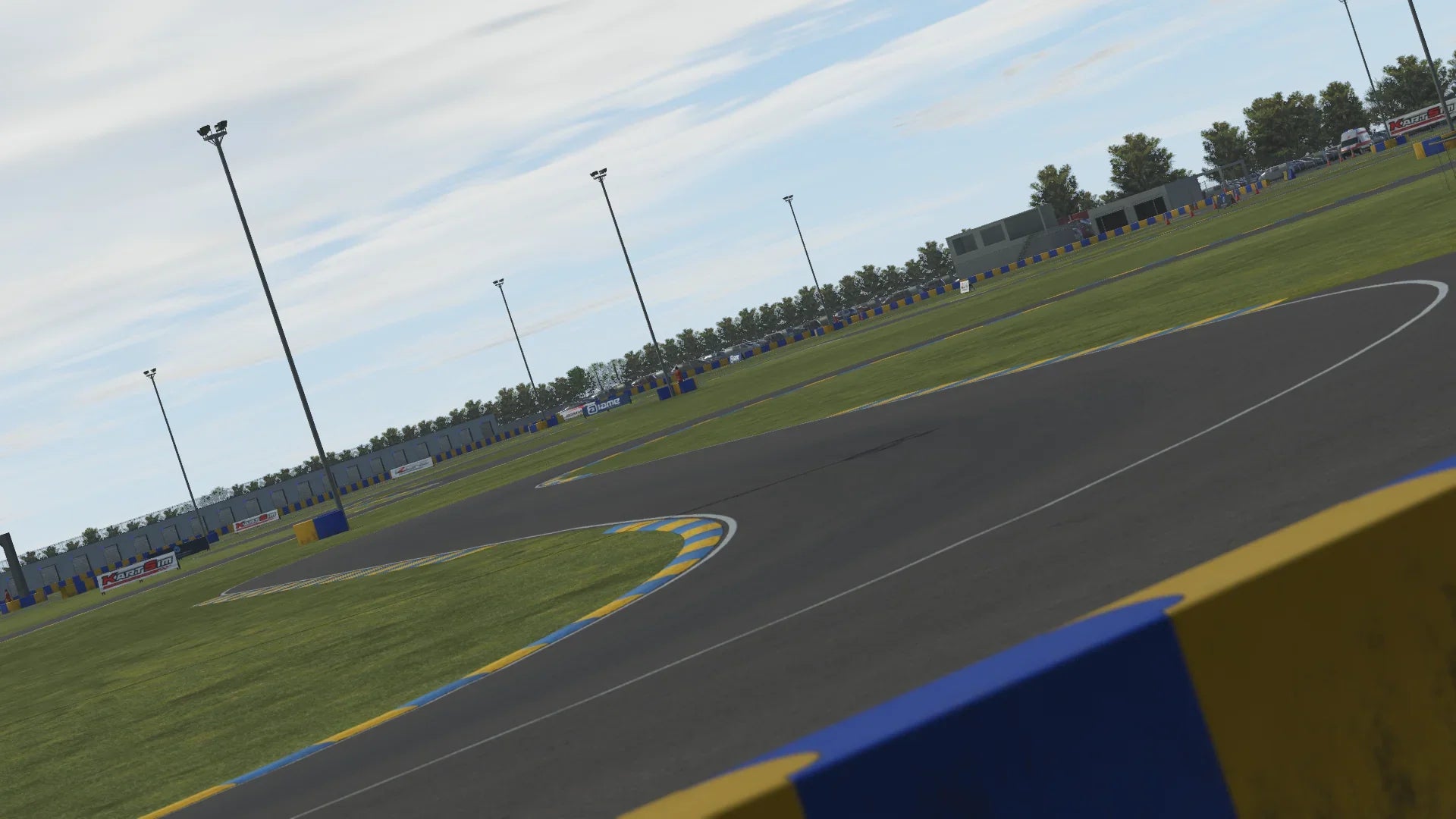 KartSim software render of a Le Mans–style kart track corner with blue-and-yellow kerbs and green grass, tall floodlight poles lining the circuit, pit building and barriers in the distance, and scattered advertising boards under a partly cloudy sky.