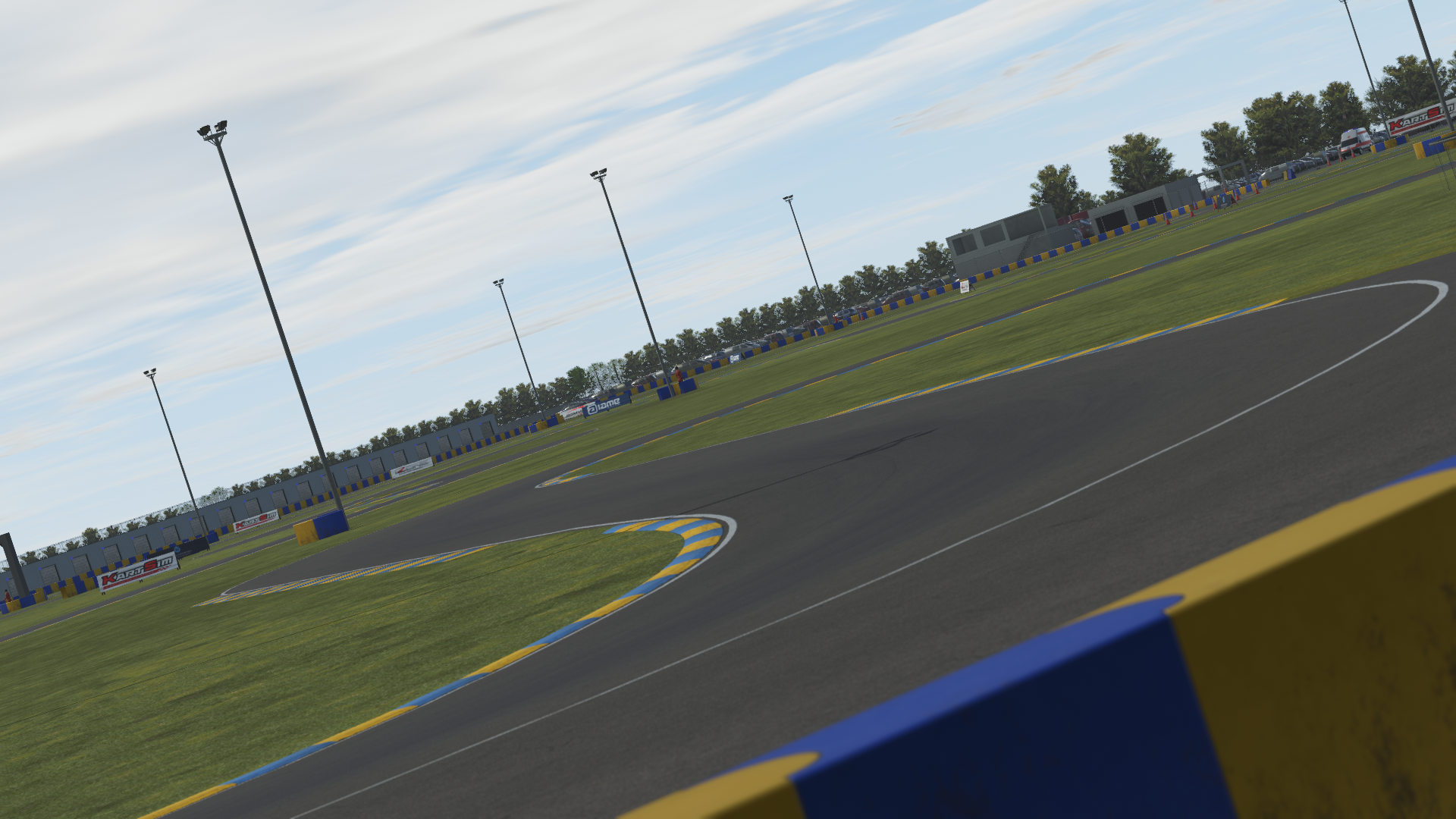 KartSim software render of a Le Mans–style kart track corner with blue-and-yellow kerbs and green grass, tall floodlight poles lining the circuit, pit building and barriers in the distance, and scattered advertising boards under a partly cloudy sky.