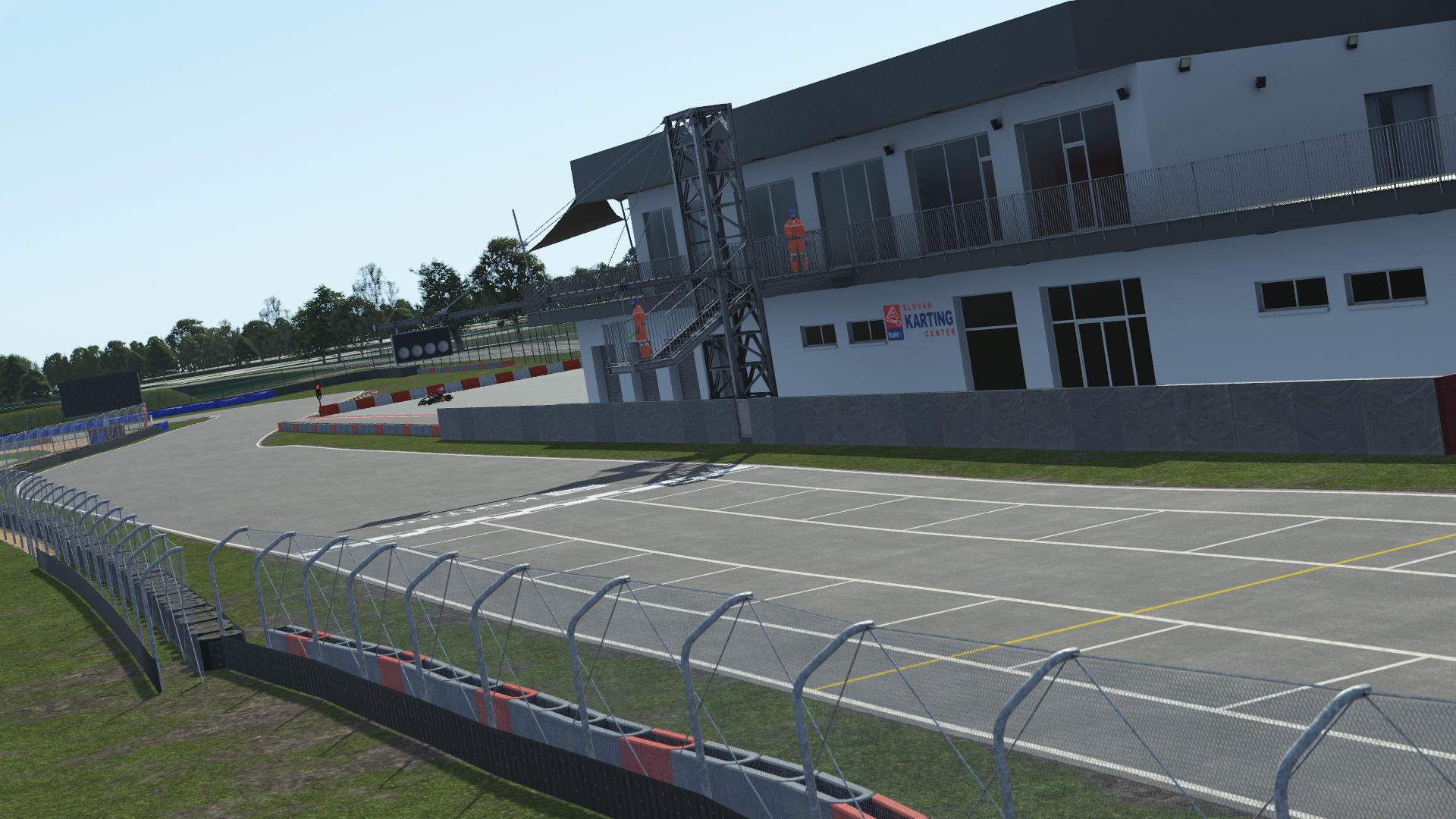 View of the pit building and start/finish straight at the Slovakiaring karting circuit in KartSim software, showing the asphalt track with red-white kerbs, protective fencing, pit garages, and clear sky above.