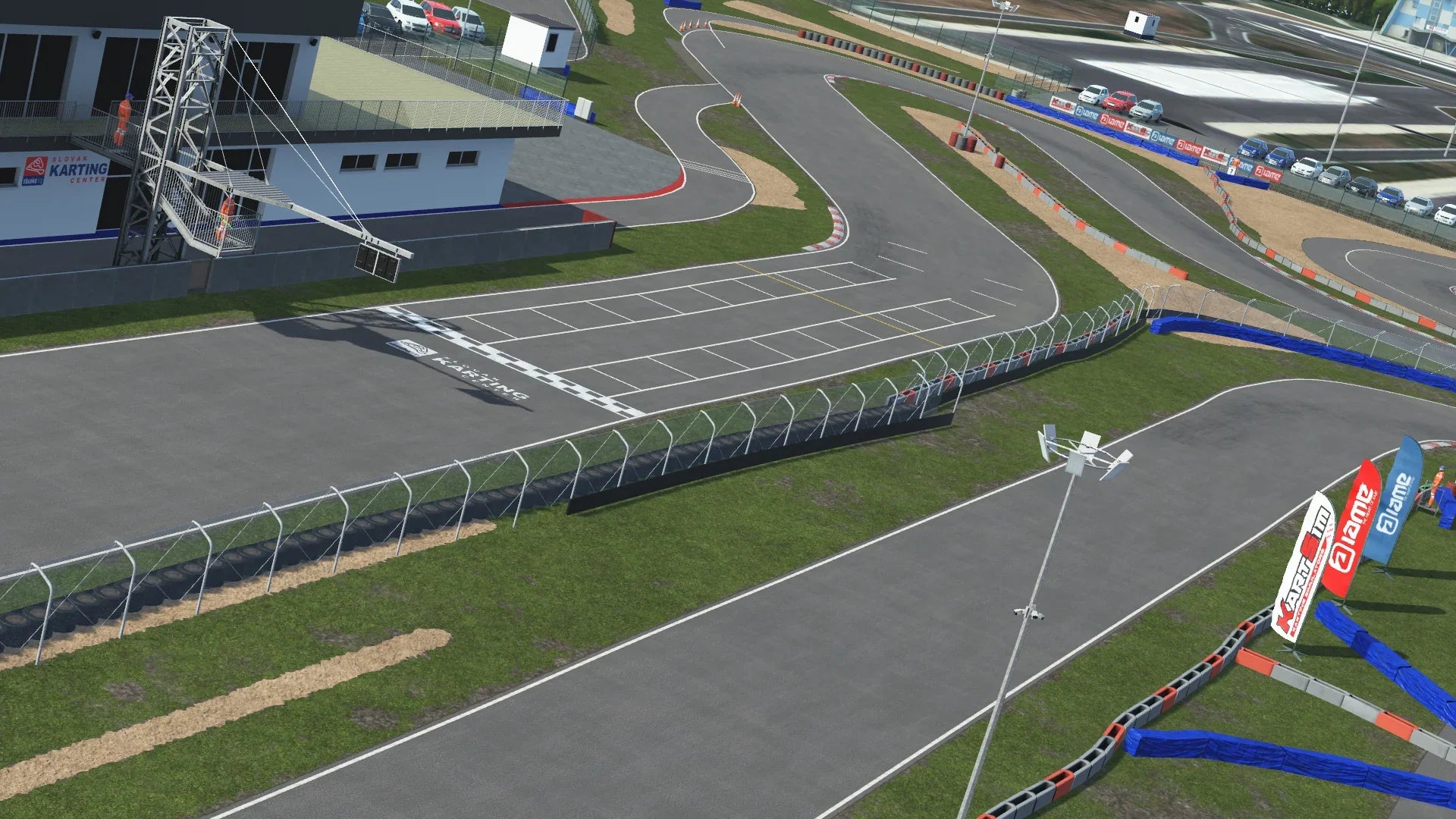 View of the pit lane and grandstand at the Slovakiaring karting circuit in KartSim software, showing the asphalt start/finish straight, red-white kerbs, protective fencing, pit buildings, paddock cars, and spectator stands under a clear sky.