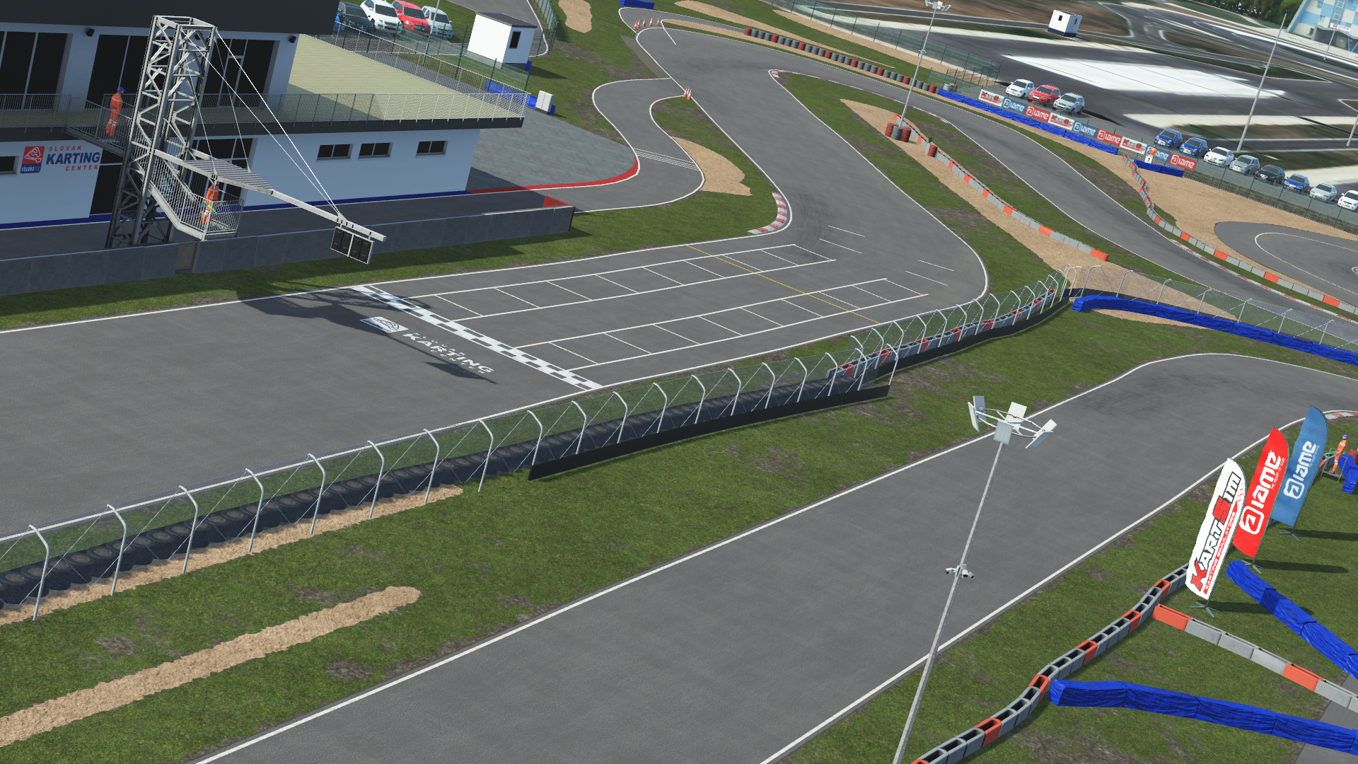 View of the pit lane and grandstand at the Slovakiaring karting circuit in KartSim software, showing the asphalt start/finish straight, red-white kerbs, protective fencing, pit buildings, paddock cars, and spectator stands under a clear sky.