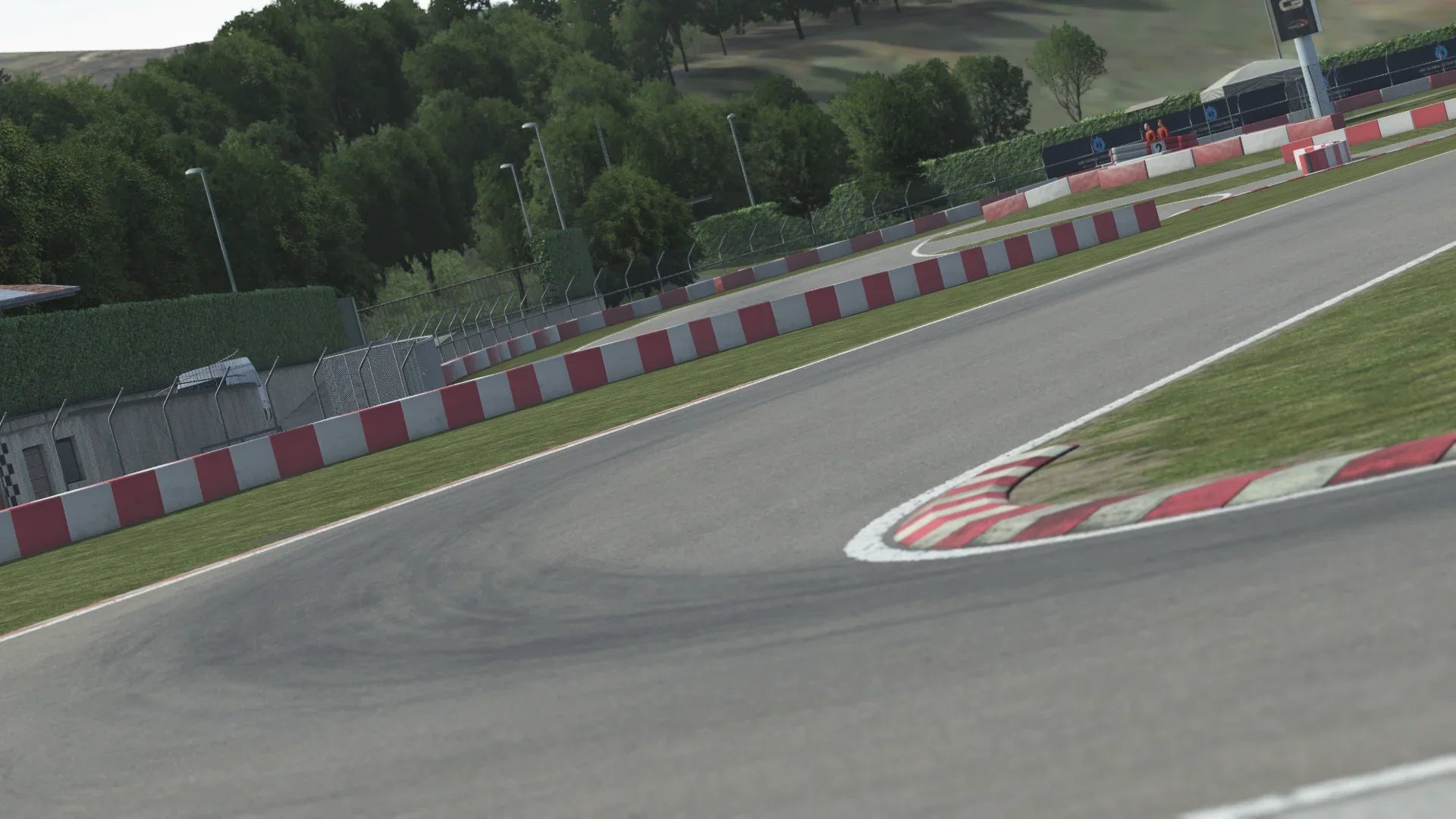KartSim software close-up of South Garda kart circuit corner, showing an asphalt turn with red-and-white curbs, green grass verge, perimeter fencing and a backdrop of trees and trackside buildings under a clear sky.