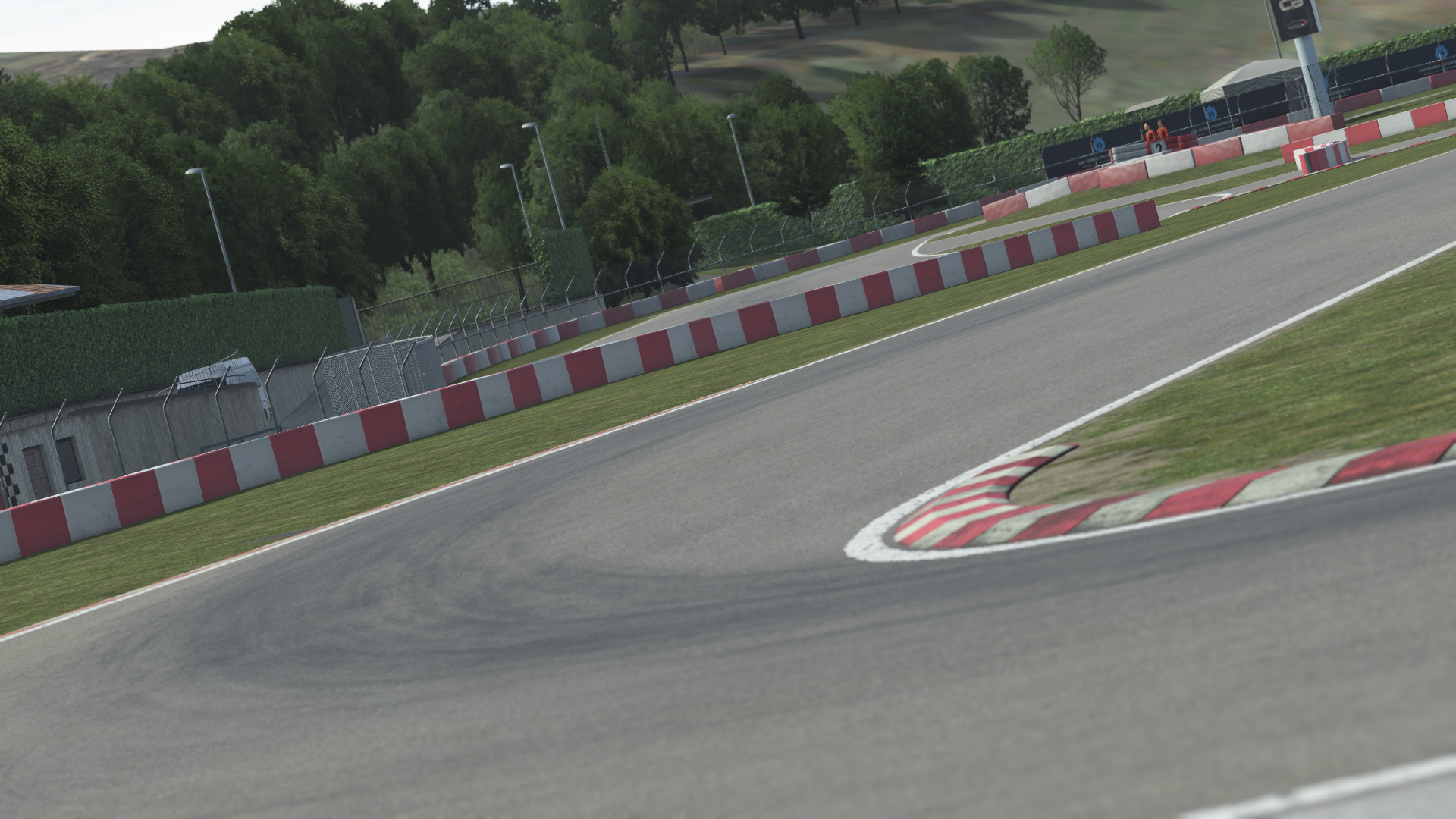 KartSim software close-up of South Garda kart circuit corner, showing an asphalt turn with red-and-white curbs, green grass verge, perimeter fencing and a backdrop of trees and trackside buildings under a clear sky.