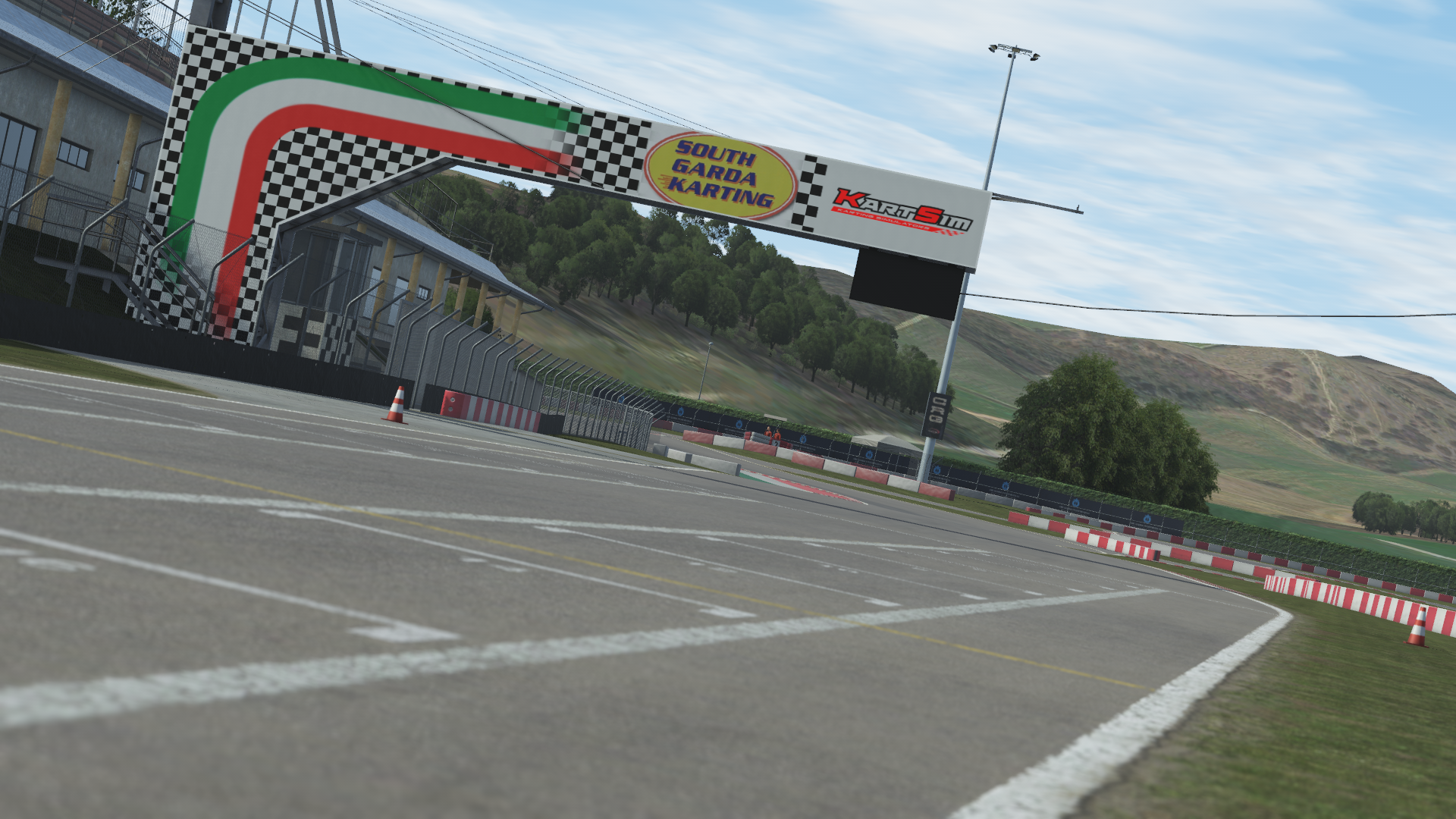 KartSim software screenshot of the South Garda Karting finish-line arch, featuring a green-white-red Italian flag motif, checkered pattern, KartSim and track logos, with the grid-lined asphalt track and surrounding hills in the background under a clear sky.