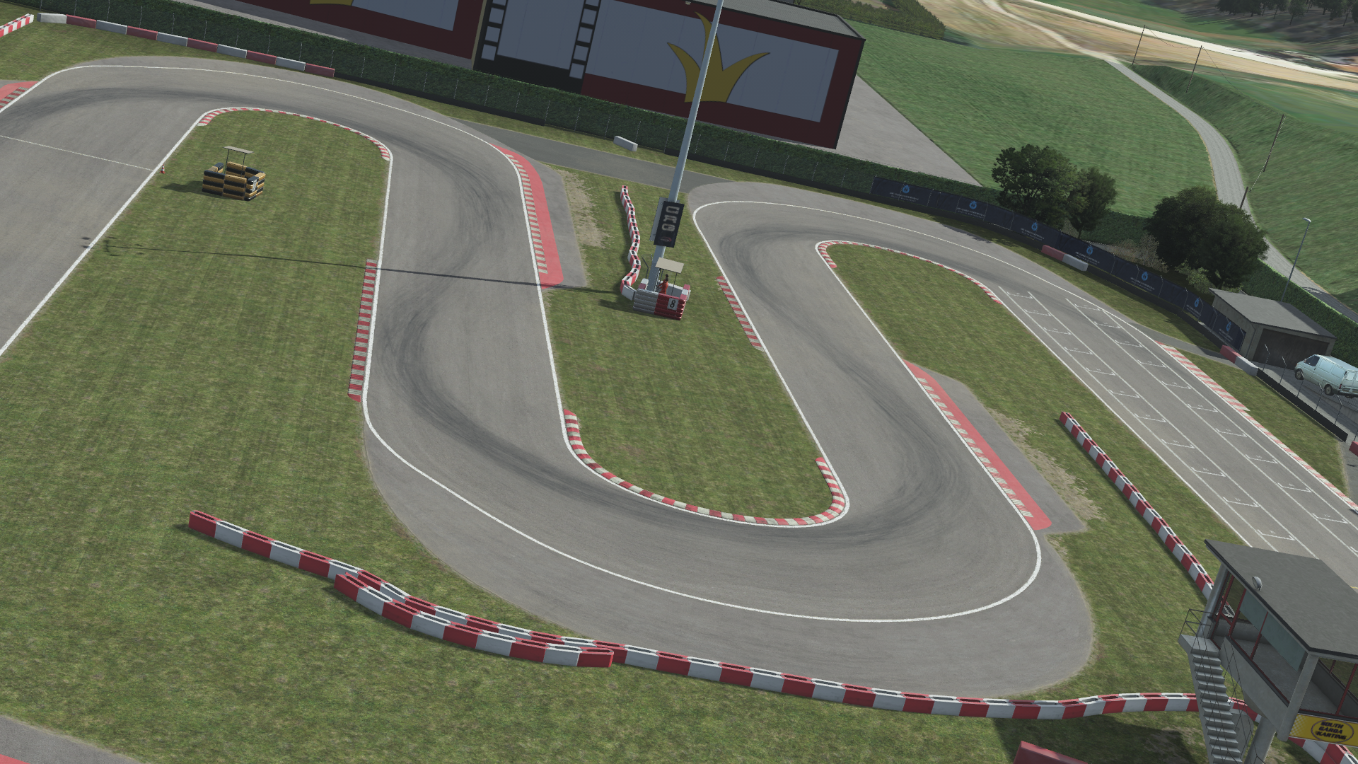 KartSim software overhead rendering of South Garda kart circuit, featuring a tight asphalt chicane lined with red-and-white barriers, lush green grass infield, a control tower and pit building at trackside, and rolling hills in the distance.