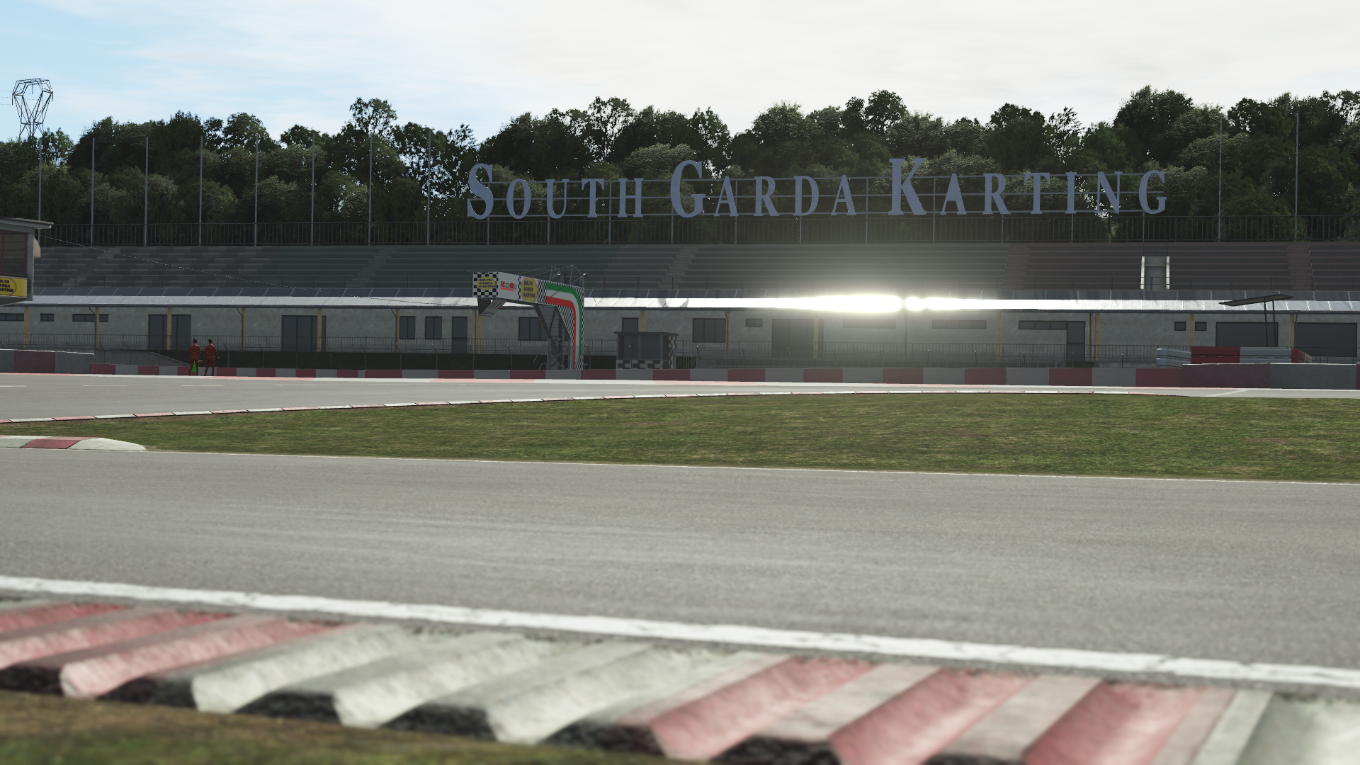 KartSim software image render showing the South Garda Karting grandstand signage above the pit building, with the asphalt track and grassy infield in the foreground under an overcast sky.