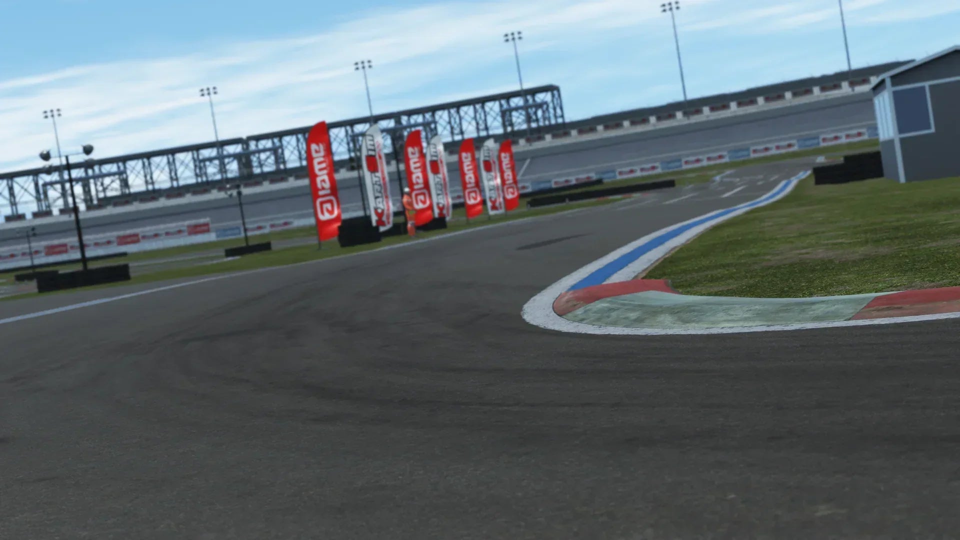 Close-up of a racetrack corner with red and white curbs, tire marks, and sponsor flags, with grandstands and a bridge in the background