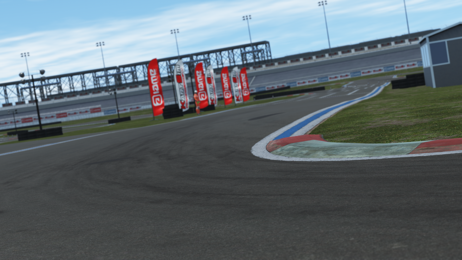 Close-up of a racetrack corner with red and white curbs, tire marks, and sponsor flags, with grandstands and a bridge in the background