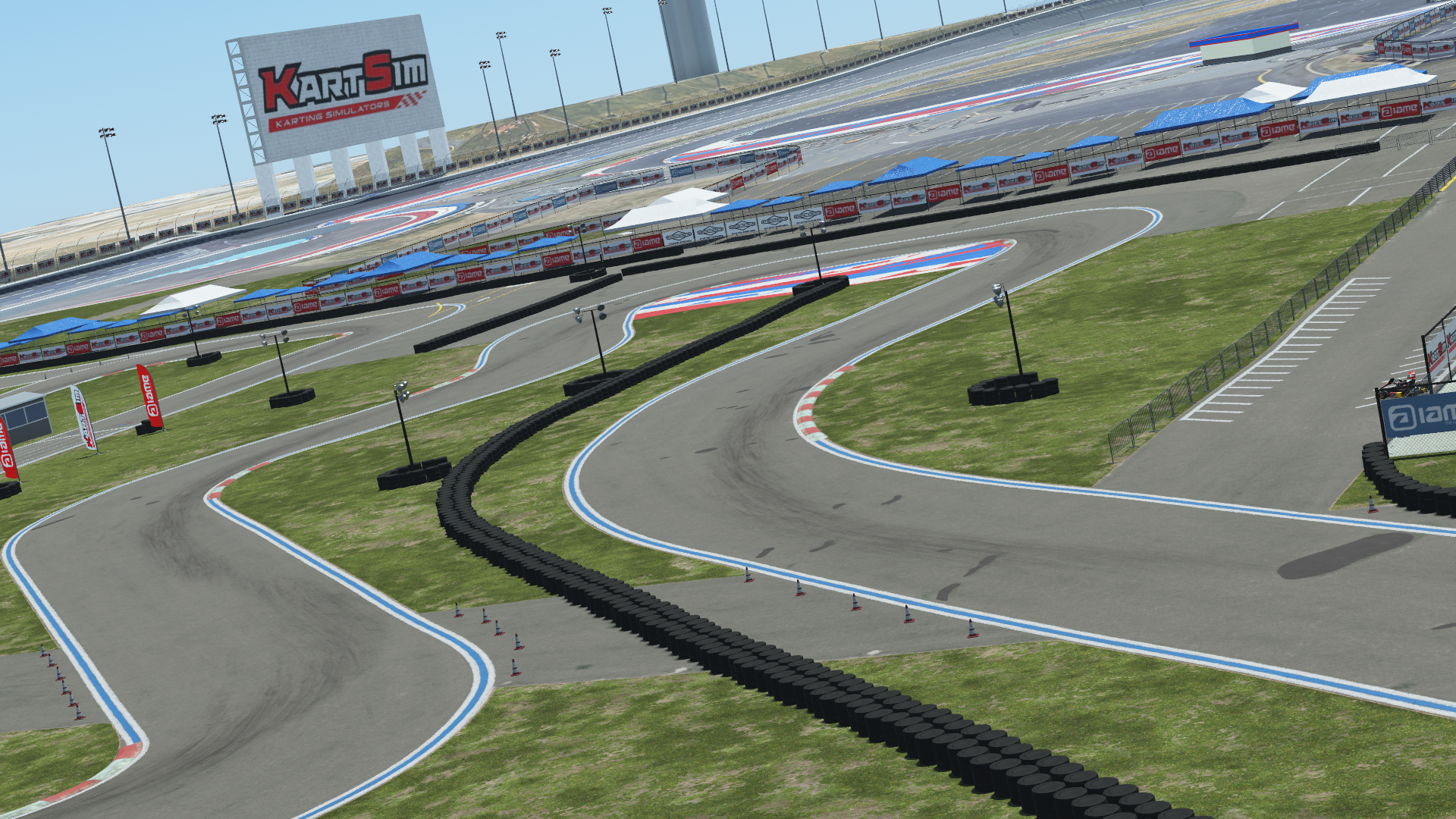 Wide view of a karting circuit with tire barriers, sponsor banners, and a large KartSim billboard under a blue sky