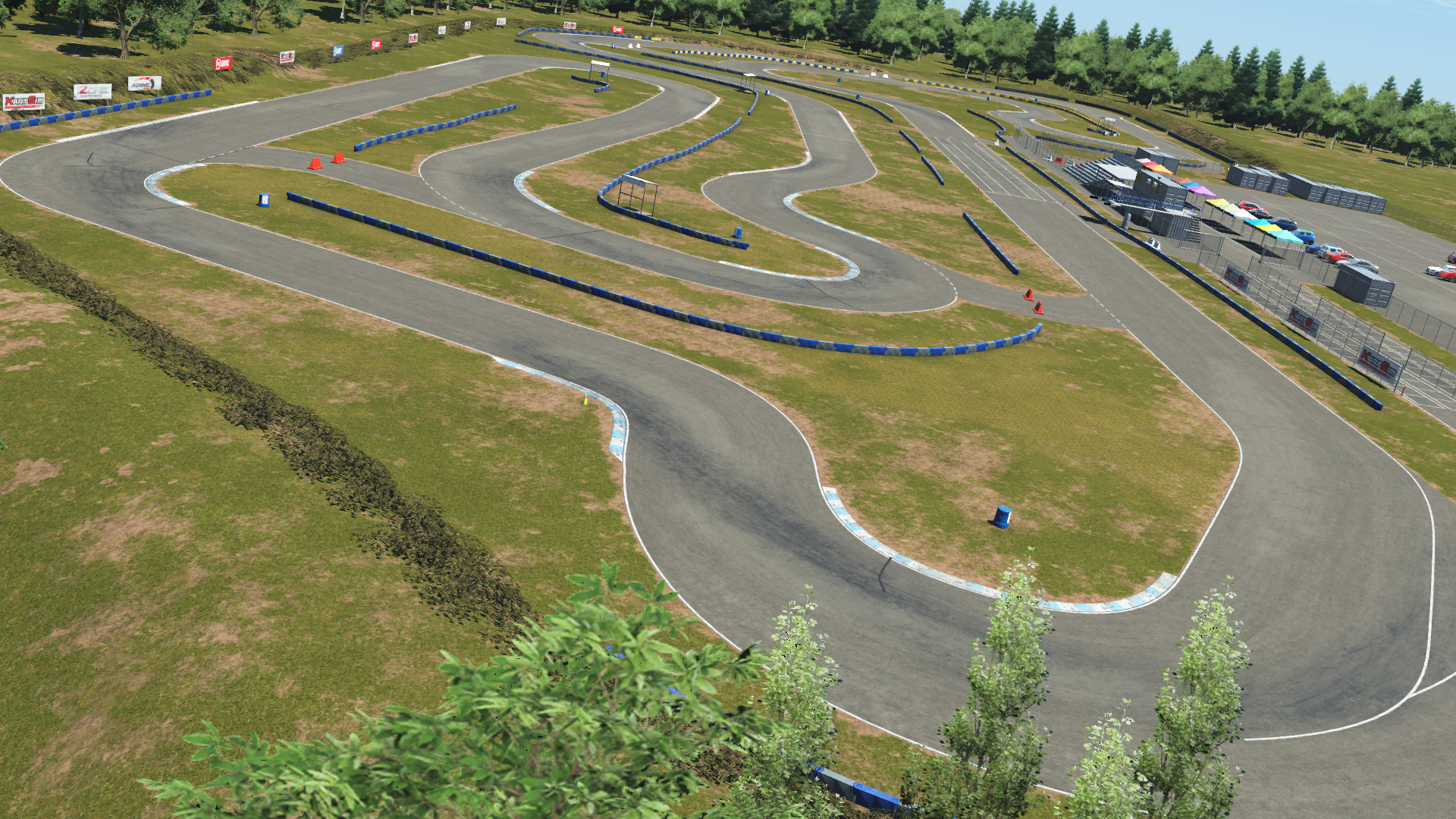 Wide aerial view of Greg Moore karting circuit with winding turns, blue barriers, grassy patches, and parked cars near the paddock area