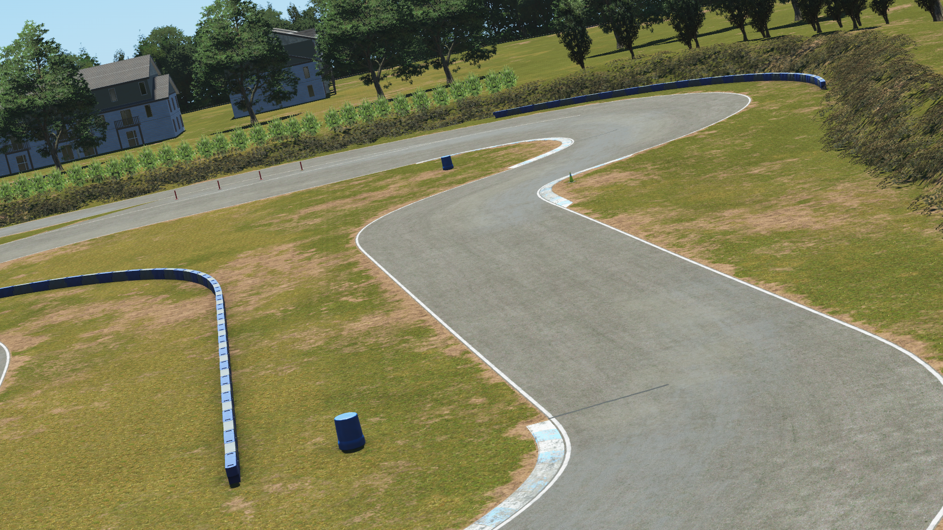 Curving section of Greg Moore kart track with sparse blue barriers, grassy surroundings, and residential houses in the background