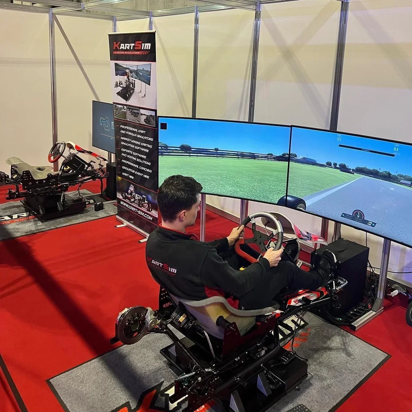 KartSim motion simulator demonstration at a trade show featuring a user driving a kart-style rig with triple screen display, red flooring, and promotional banners in the background