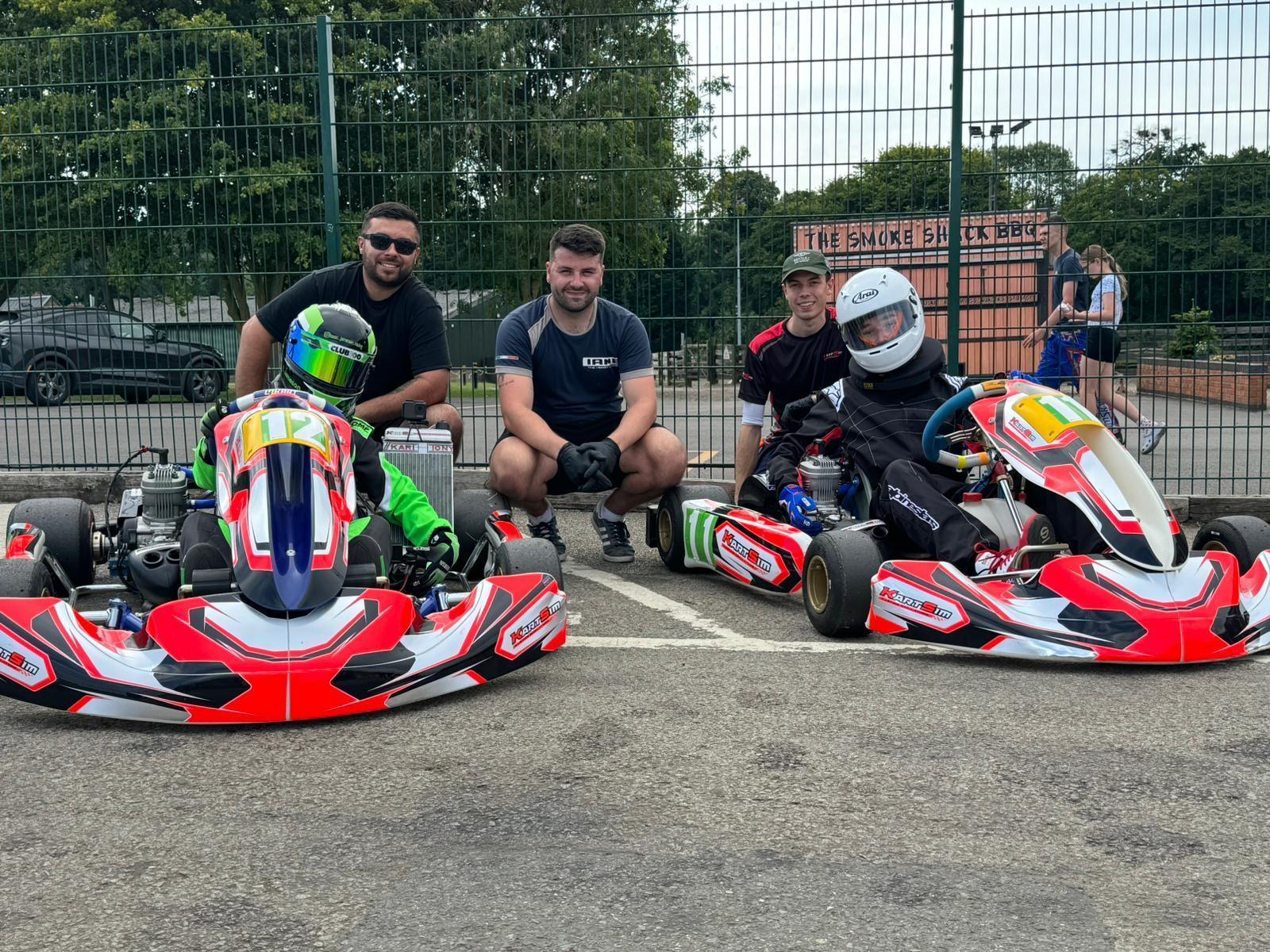 KartSim trackside kart hire package featuring two drivers in full gear with race karts, accompanied by support team in paddock environment