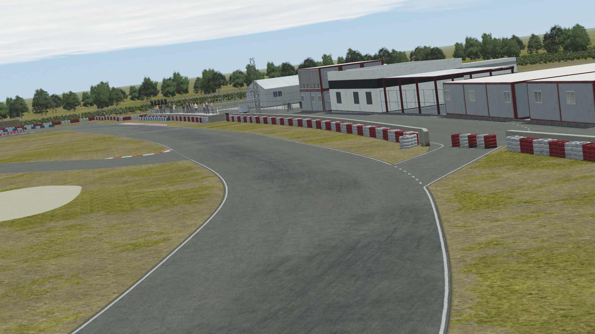 In-simulation view of the Valencia karting circuit pit complex in KartSim software, showing the asphalt track with red-white barriers, pit buildings, control tower, surrounding trees, and a partly cloudy sky.