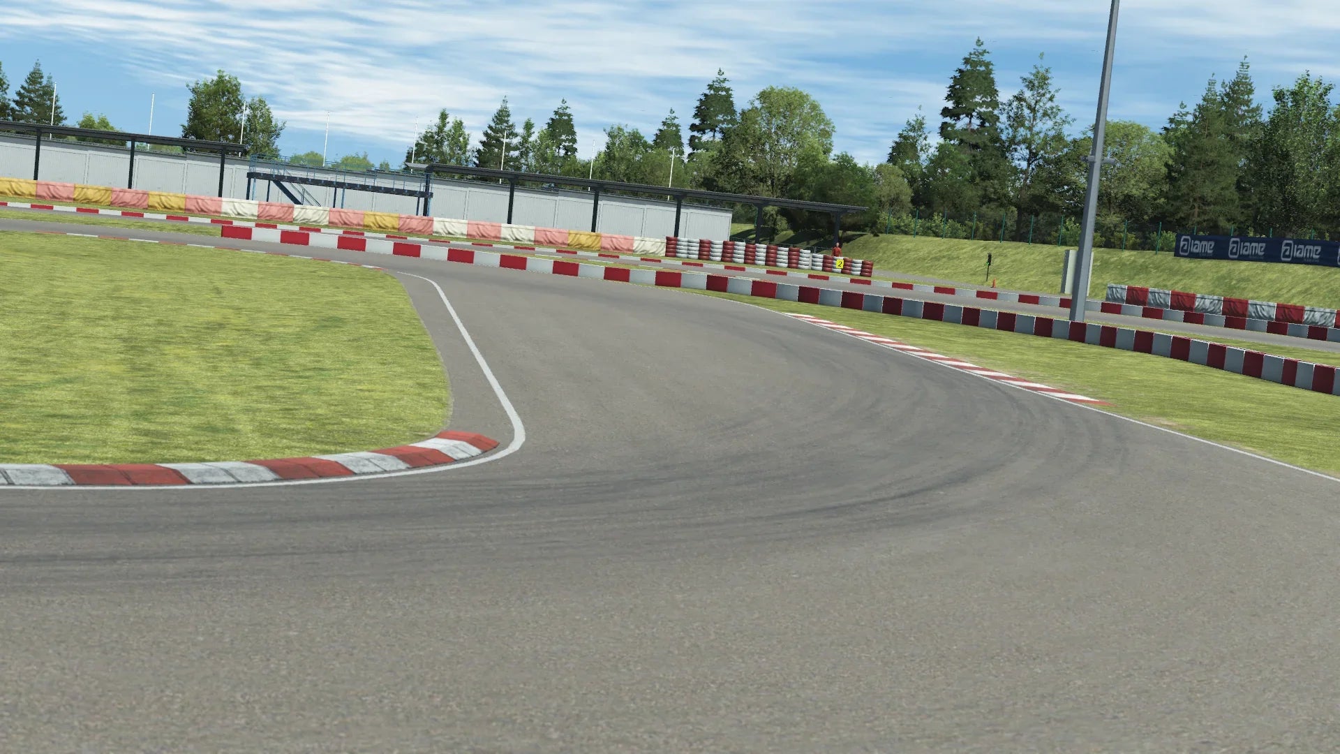 Curved in-simulation view of the Wackersdorf karting circuit in KartSim software, featuring the asphalt corner with red-white kerbs, grass run-off area, protective barriers, pit buildings, and surrounding forest under a clear sky.