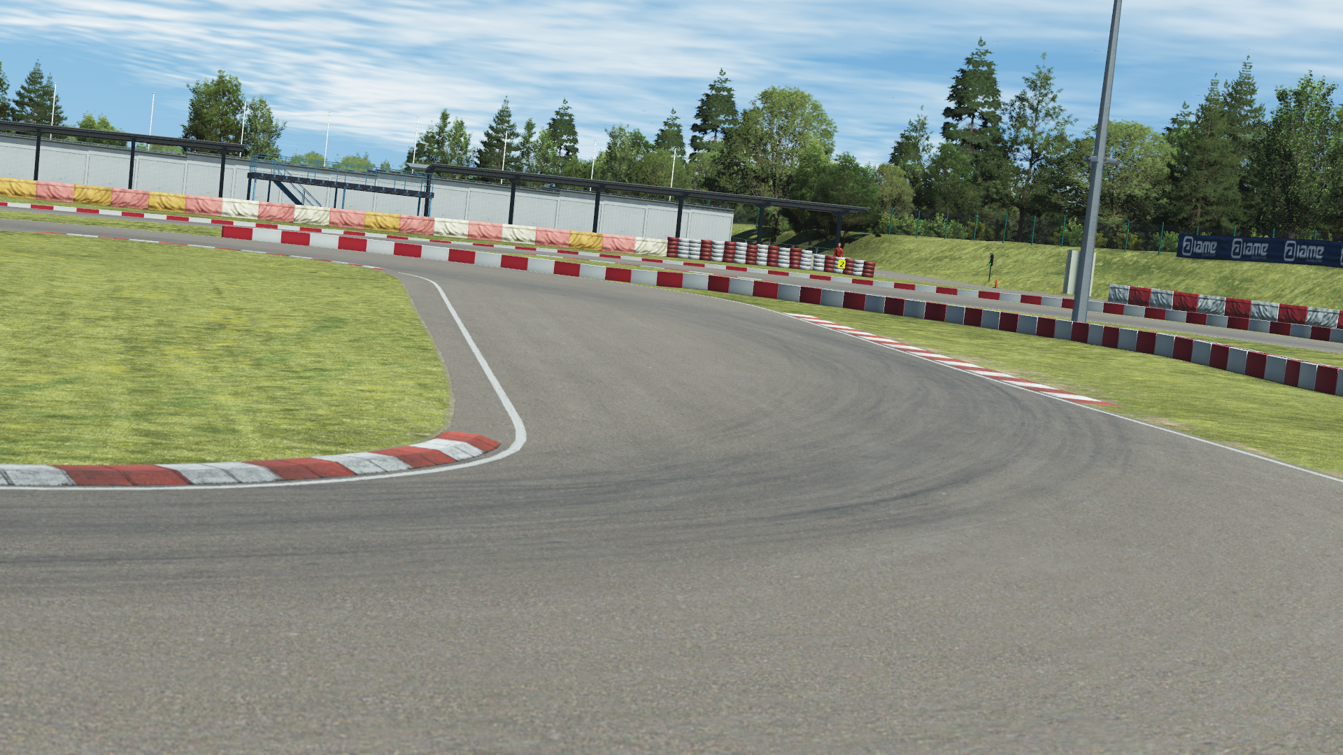 Curved in-simulation view of the Wackersdorf karting circuit in KartSim software, featuring the asphalt corner with red-white kerbs, grass run-off area, protective barriers, pit buildings, and surrounding forest under a clear sky.