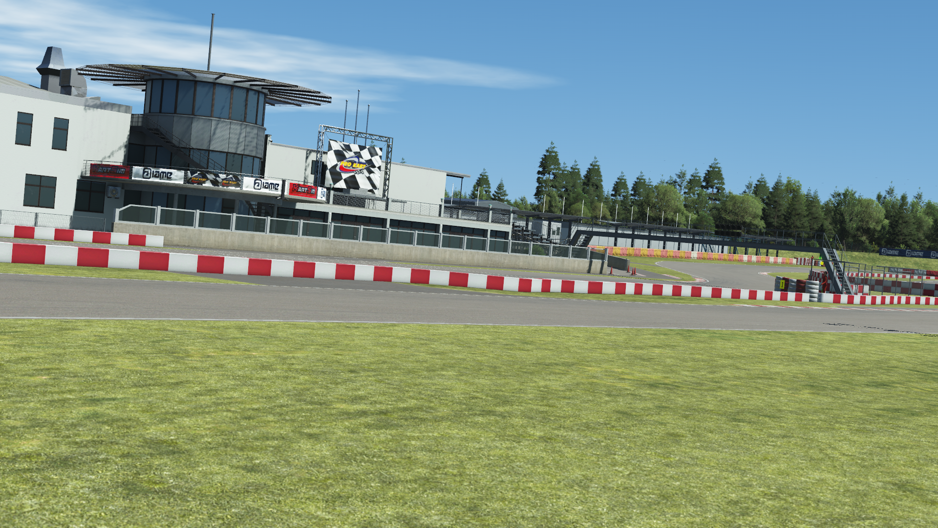 In-simulation view of the pit building and control tower at the Wackersdorf karting circuit in KartSim software, showing the asphalt track with red-white barriers, grass run-off areas, pit grandstand, and clear blue sky.