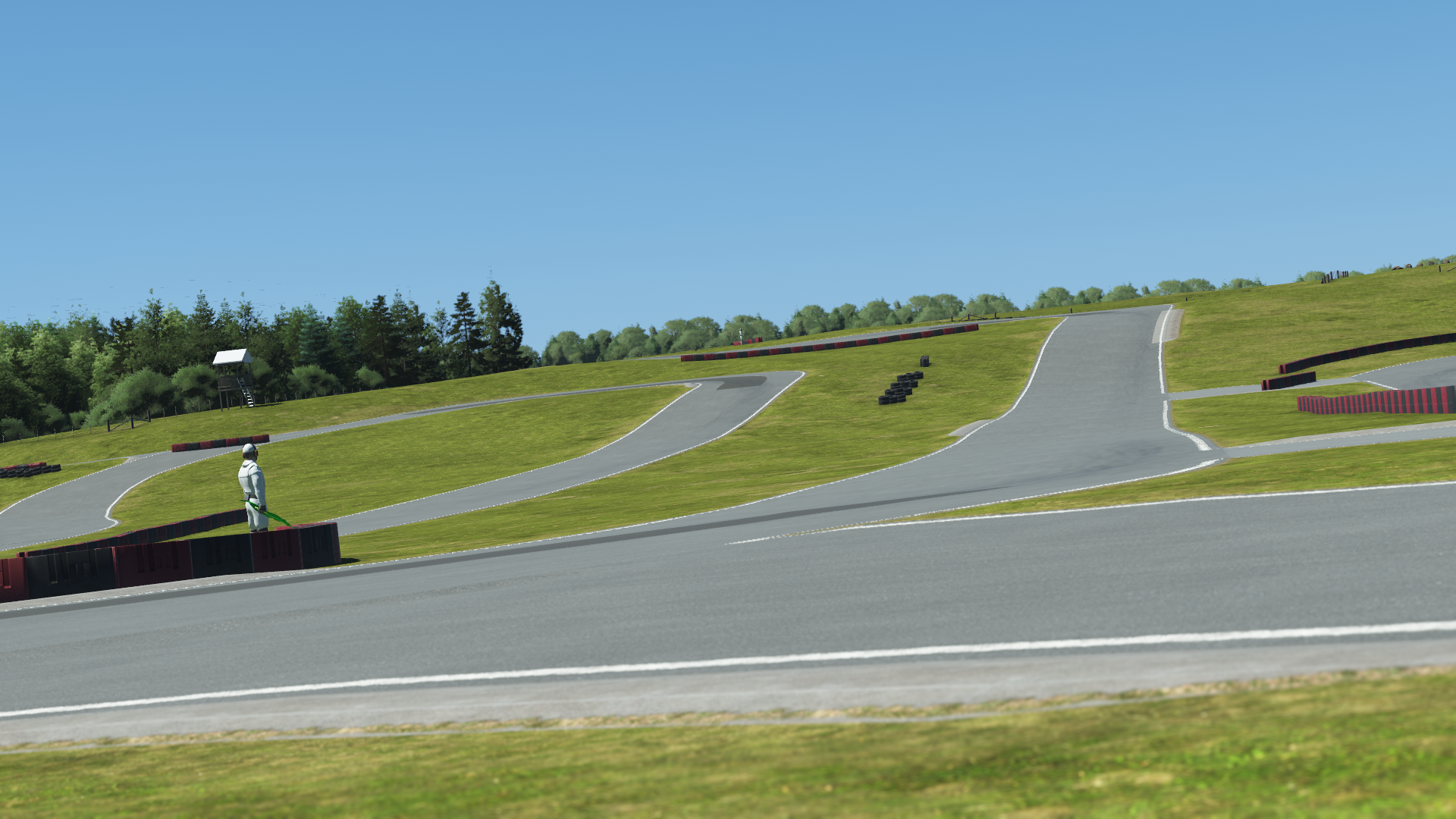 Elevated section of Mosport karting track with steep incline, red and black barriers, grassy slopes, and tree line under clear sky